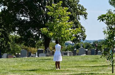 A woman stands in a cemetery tying a prayer ribbon to a small tree. Rows of graves are in the background.