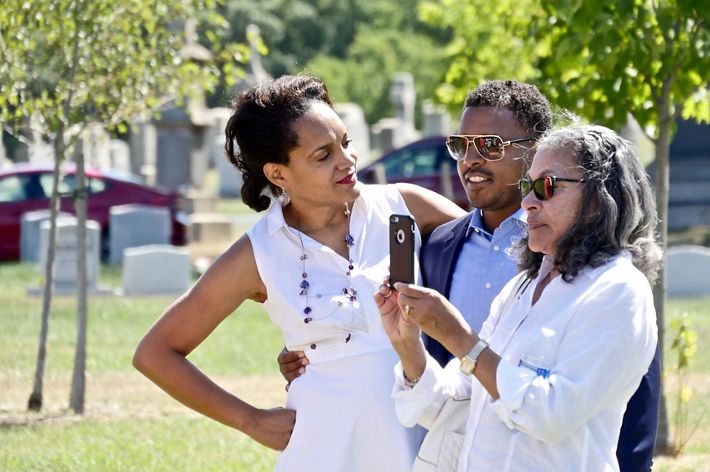 Three people stand together during a family gathering. One of the people uses her cell phone to take a picture of someone out of view.