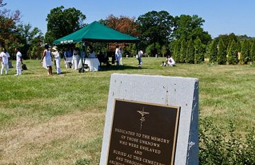 A group of people gather together under a tent during a family gathering. A large granite memorial in the foreground honors the enslaved and unknown individuals buried at the