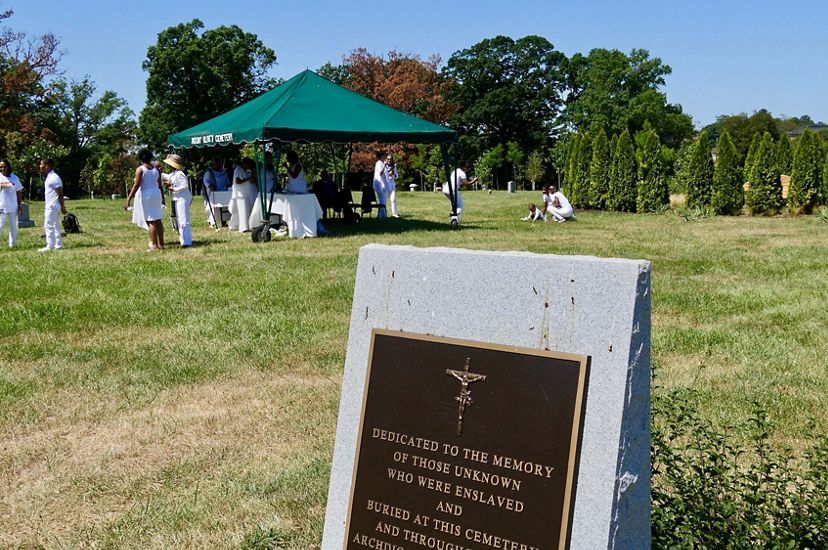 A group of people gather together under a tent during a family gathering. A large granite memorial in the foreground honors the enslaved and unknown individuals buried at the site.