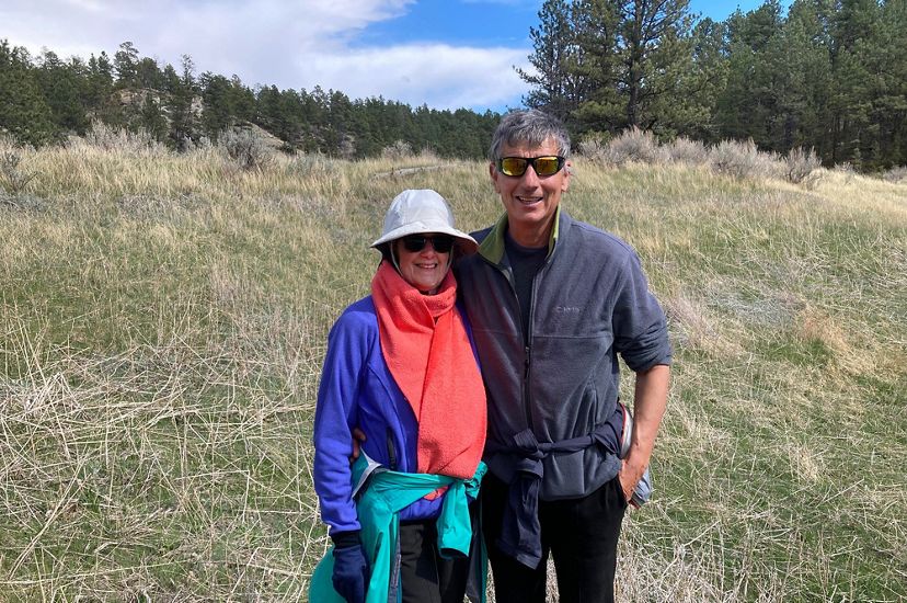 Kate and Curt Vogel standing on a grassy slope with a forested ridge in the background.