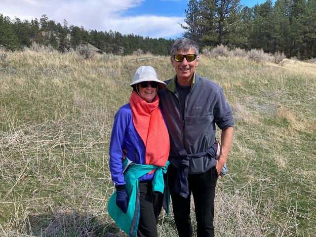 Kate and Curt Vogel standing on a grassy slope with a forested ridge in the background.