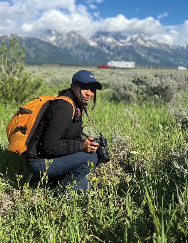 Mars Flores squats near the ground with a camera in hand, with mountains in the distance.