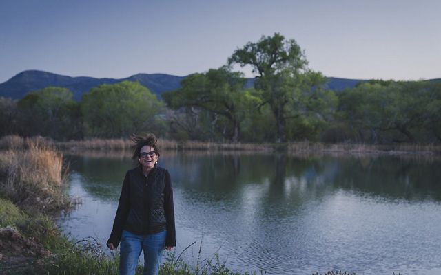 A smiling woman stands next to a large pond. The rippled surface of the water reflects a lone tree growing on the bank and mountains in the distance.