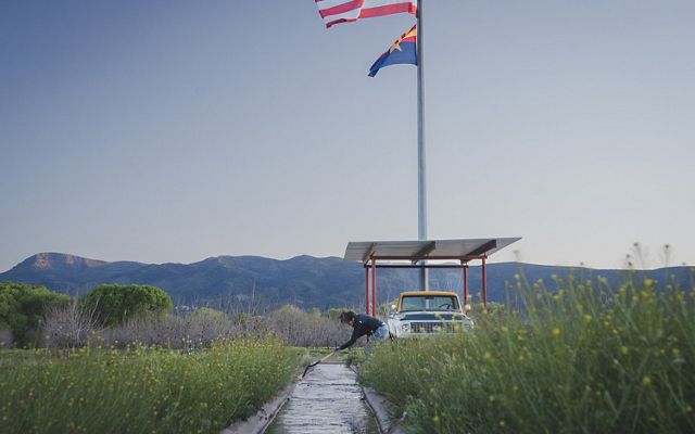 A woman stands at the edge of an irrigation ditch using a shovel to clear away debris. A flagpole rises behind her and tall, flowering grasses grow all around.