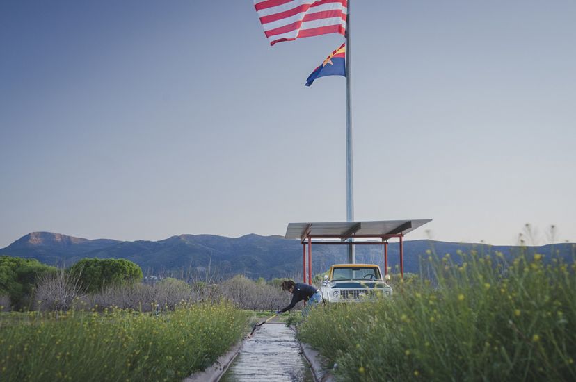 A woman stands at the edge of an irrigation ditch using a shovel to clear away debris. A flagpole rises behind her and tall, flowering grasses grow all around.