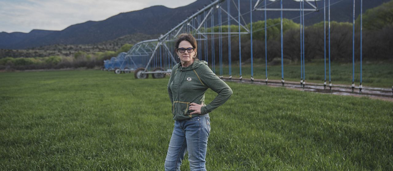 Claudia Hauser, wearing sunglasses, jeans and a green hoodie, stands with her hands on her hips in a verdant field of young grass, with her center pivot irrigation structure behind her. 