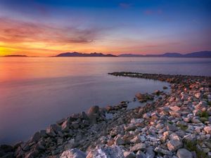 The sun sets over the Great Salt Lake, with a jetty of rocks curving into the water in the foreground.