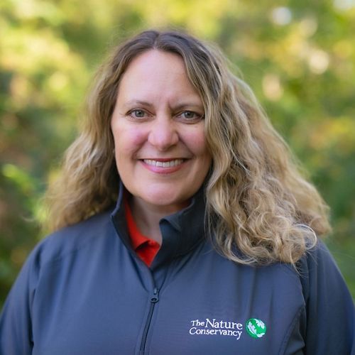 A headshot of a woman with blonde curly hair, wearing a jacket that says The Nature Conservancy, standing outside.