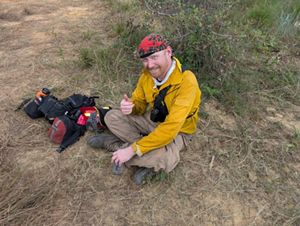 John Lerdal sits on the ground and gives a thumbs up to the camera.