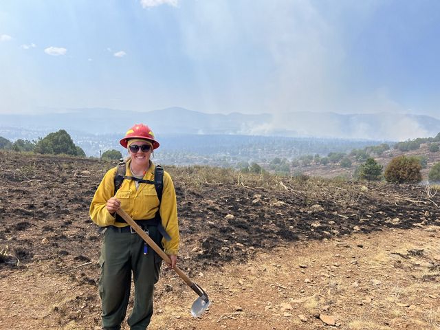 A woman wearing a yellow fire jacket, black pants, and a red helmet holds fire ax while standing outside with mountains in the background.
