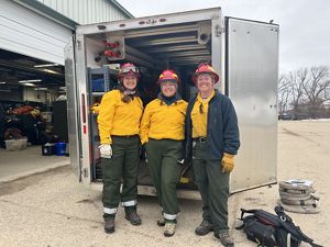 Three women dressed in fire gear standing next to fire equipment. 