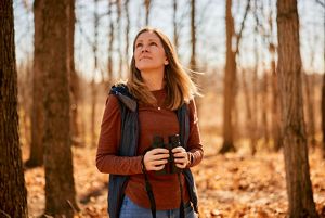 A woman standing in a forest with binoculars. 