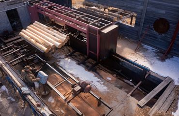 Overhead view of Paul Rust standing at a computer attached to sawmill machinery that has large logs on it.
