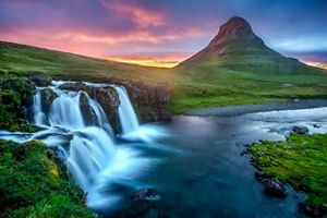 Water rushes over a waterfall in a green landscape with a large pointed hill in the background and a vibrant orange sunset peeking around the hill.