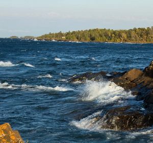 Waves crash against rocky shoreline rocks on a large lake, sending white spray into the air. A sunlit forested peninsula stretches across the background beneath a pale blue sky.