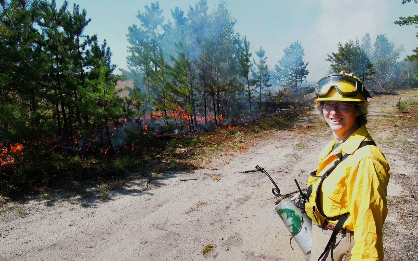 A woman holding as drip torch in a yellow fire protective suit stands with a fire burning behind.