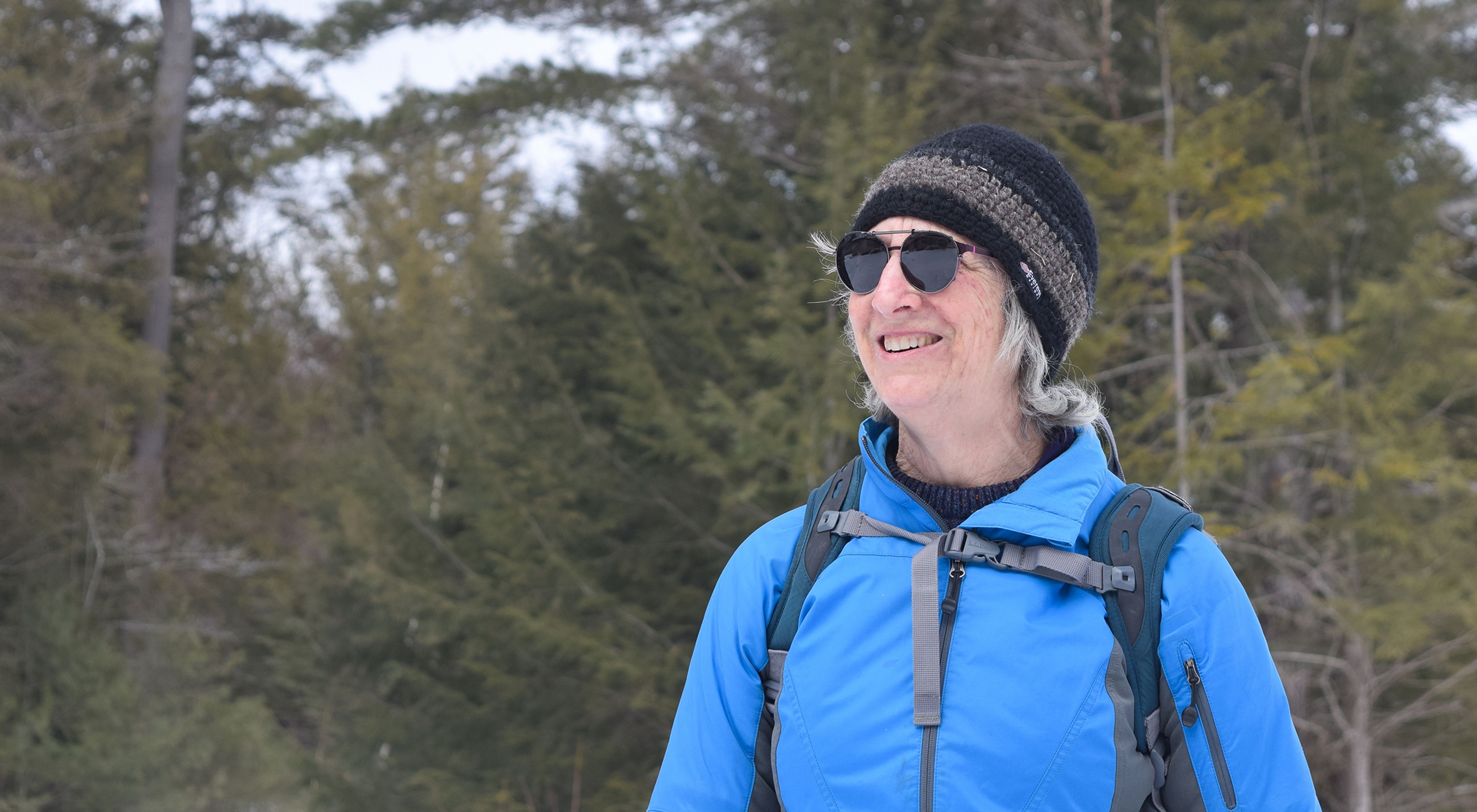 A woman in dark sunglasses, wool hat and blue jacket smiles with evergreen trees behind her.