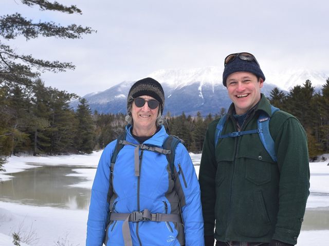 A woman and man stand smiling in a wintry scene with a mountain behind them.