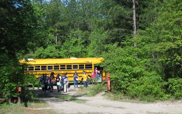 Middle school students file out of a school bus, walking along a dirt road through a wide opening in a line of trees.
