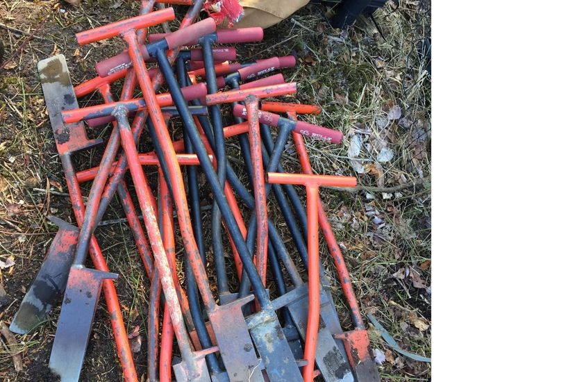 Orange handled dibble bars lay in a pile on the ground. The shovel-like tools have wide, thin blades to cut into the ground and prepare a hole for tree seedlings.