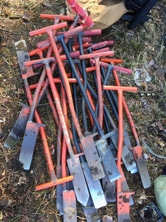 Orange handled dibble bars lay in a pile on the ground. The shovel-like tools have wide, thin blades to cut into the ground and prepare a hole for tree seedlings.