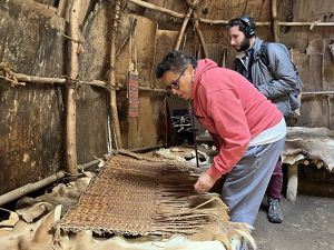 Podcast producer Jay Feinstein stands with Paula Peters of Native Land Conservancy looking at a mat of woven bulrush laid on the bench inside a Wampanoag wetu.