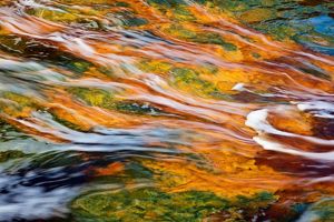 An aerial view features clear waters of a river flowing across red and orange colored rocks. 