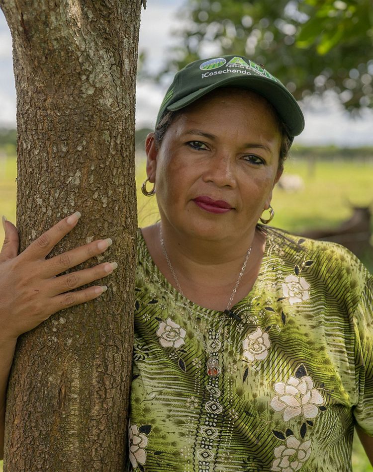 a smiling woman in a hat leans up against a small tree