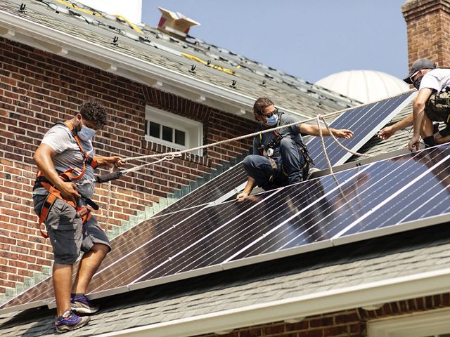 Three workers wearing masks up on a roof installing solar panels. Hilario Minaya is on the left, standing on the roof with a harness and rope for safety.