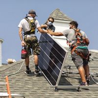 Three masked workers, viewed at center, on top of a roof installing a solar panel.