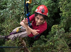 A woman looks up at the camera as she hugs a climbing rope, high up amidst the forest canopy.