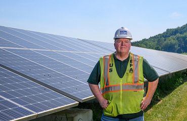 Man wearing yellow work vest stands in front of solar panels.