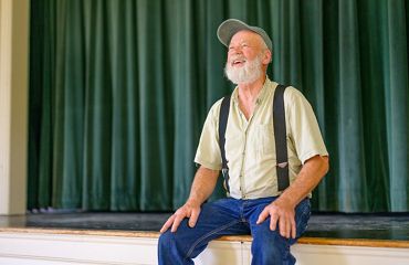 A smiling man sits on the edge of a stage. A green curtain hangs behind him.