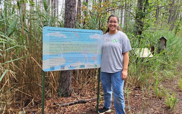 A woman wearing a Nature Conservancy tee-shirt stands next to a sign.