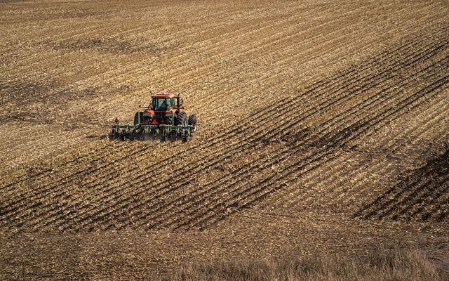 A red tractor drives through an agricultural field of golden plant remains, creating neat rows in the earth.