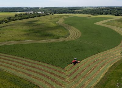 Aerial view of a red tractor cutting long, curving rows into a broad green field.