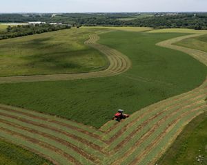 Aerial view of a red tractor cutting long, curving rows into a broad green field.