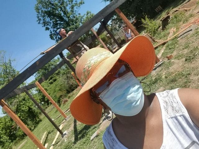 A woman in an orange straw hat, mask and sunglasses stands in front of an urban farming project.