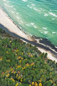 An aerial image shows a dense forested landscape and a dark river that meet a sandy beach and brightly colored lake waves.