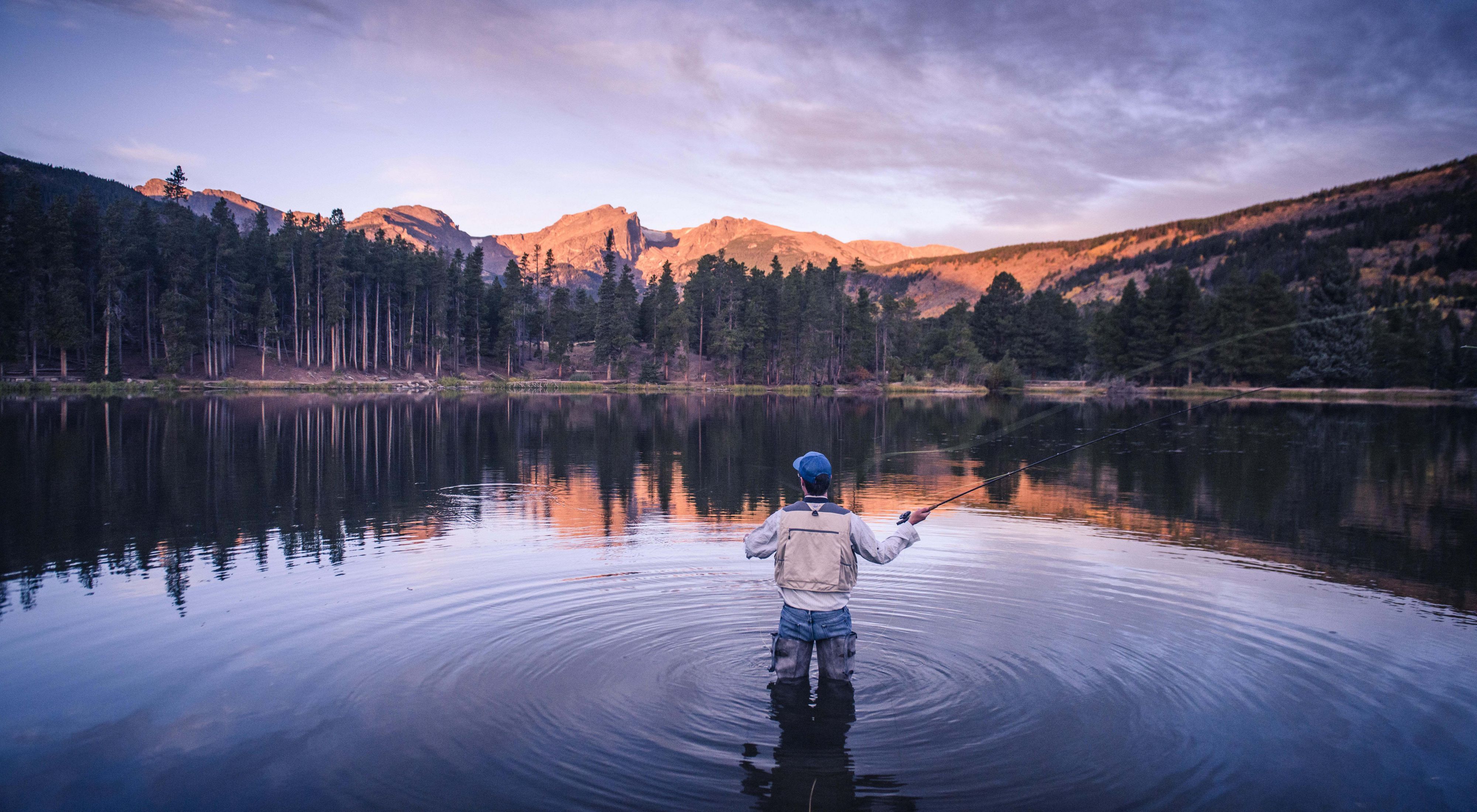 Photo of a man flyfishing in a pond in Rocky Mountain National Park, mountains in background.