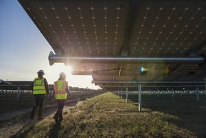 Two people wearing safety vests and hard hats walk among a field of solar panels.