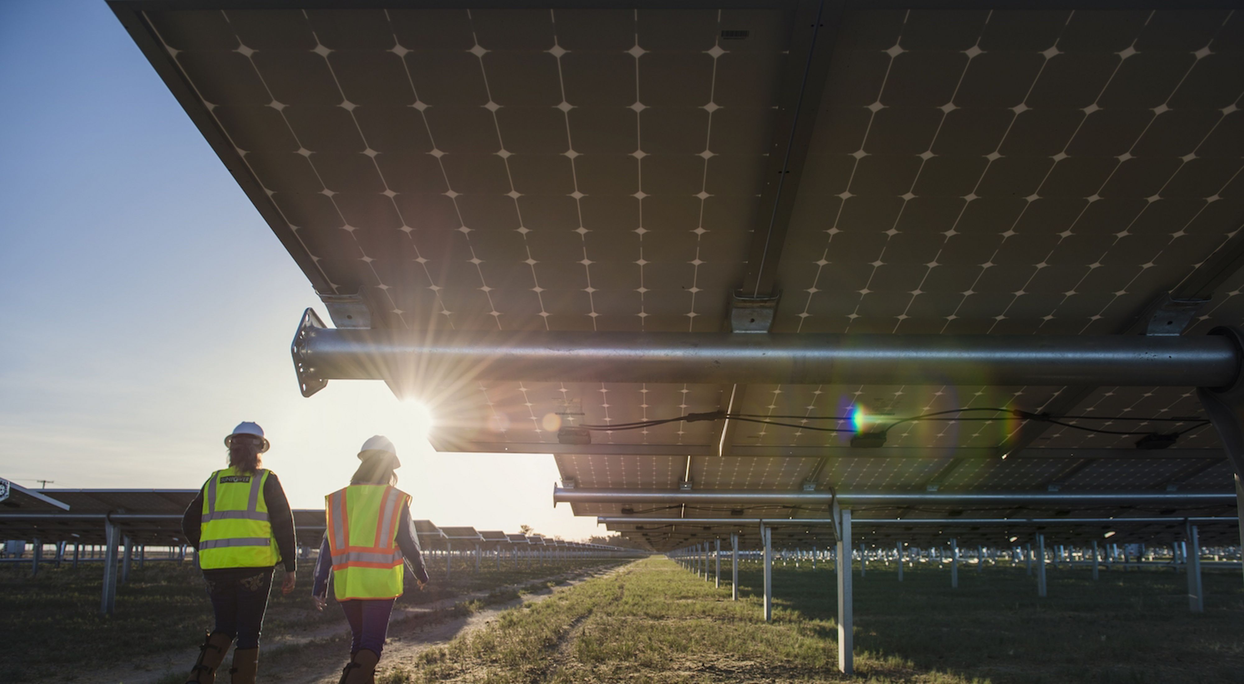 Two workers in hard hats and yellow jackets walk beneath solar panels with the sun peeking out.