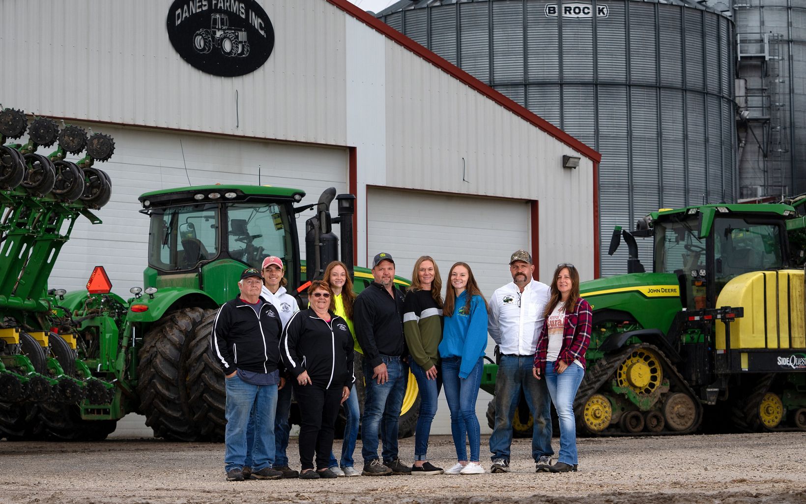 Nine members of the Danes family stand and pose in front of green tractors and metal buildings of Danes Farm.