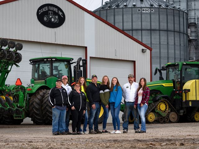 Nine members of the Danes family stand and pose in front of green tractors and metal buildings of Danes Farm.
