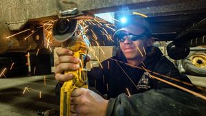 Closeup of Dylan Danes wearing protective glasses as he uses a metal grinder on a piece of machinery; sparks fly from the grinder.