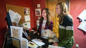 Christina Danes and Kelly Danes stand in an office room and work on a computer; their fluffy beige dog stands with its paws on the desktop.