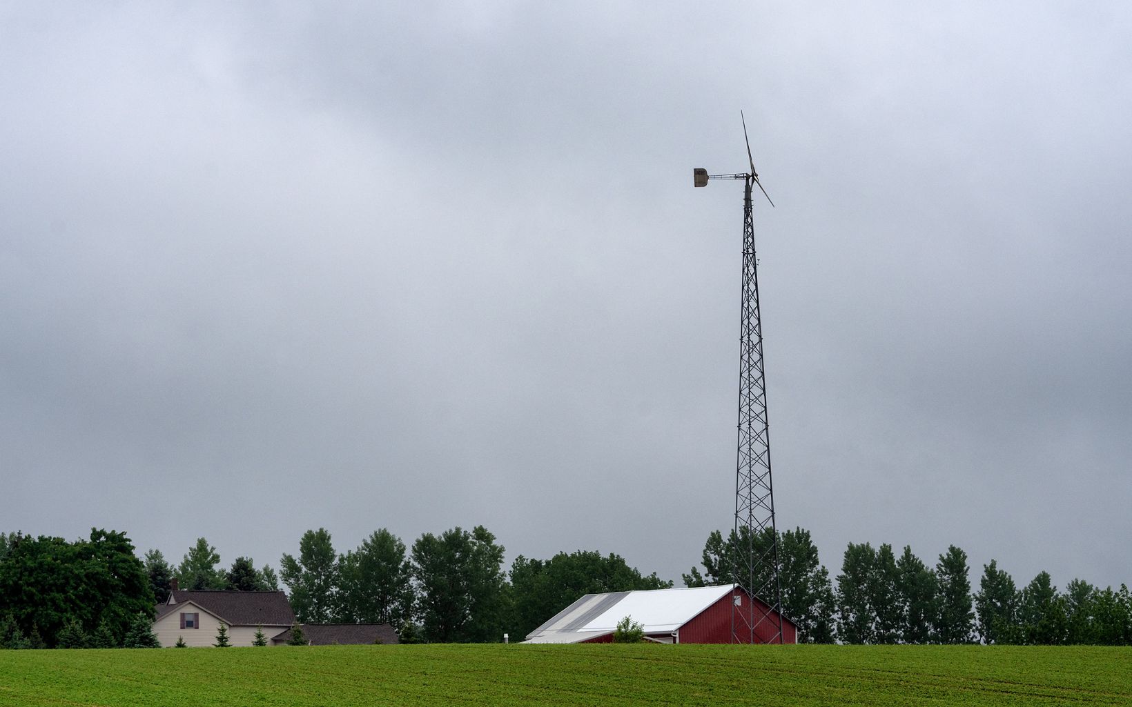 Landscape view of a steel windmill next to a red farm building and a tan house, with a row of trees in the background.
