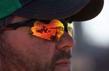 Closeup of Travis Luedke's face; he wears reflective wraparound sunglasses with orange lenses that show two farm tractors that he's looking at.