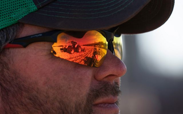 Closeup of Travis Luedke's face; he wears reflective wraparound sunglasses with orange lenses that show two farm tractors that he's looking at.